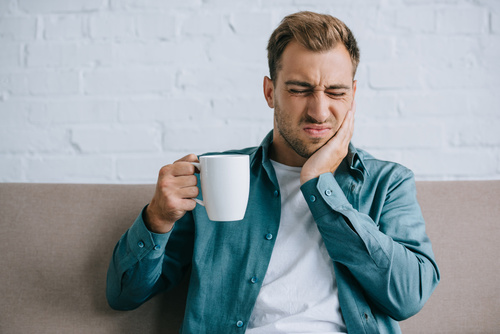 Junger Mann mit einer Tasse hält bei schmerzerfülltem Gesichtsausdruck eine Hand an die Wange (Aphthen)